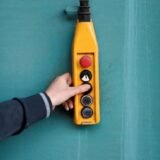 Close-up of a hand pressing a button on a yellow industrial control panel against a blue wall.